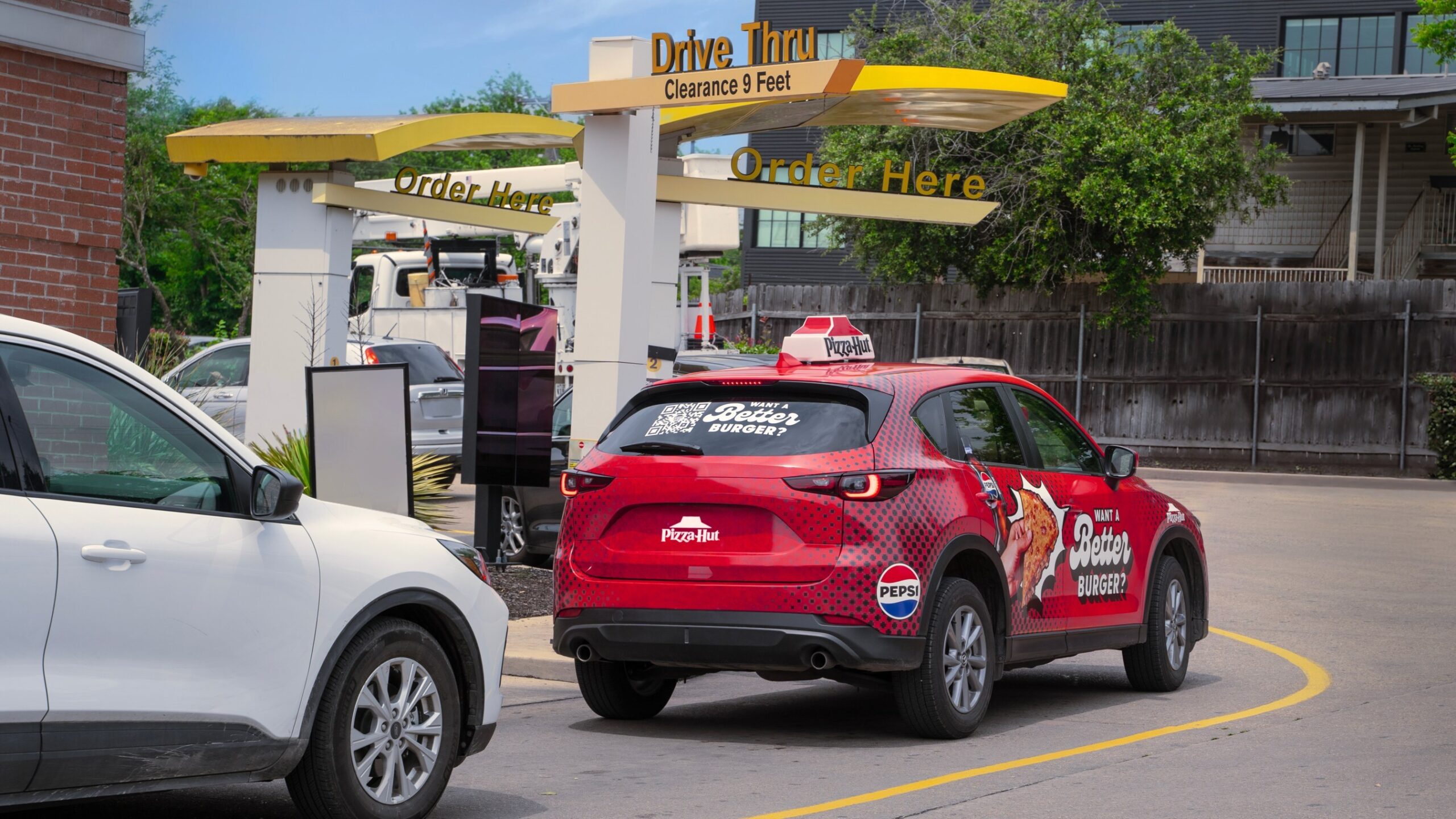 "Image: Pizza Hut delivery vehicles parked in a fast food drive-thru, displaying a QR code on their back windows to promote the new Cheeseburger Melt. Customers can scan the code to receive a coupon for a free burger and Pepsi drink."