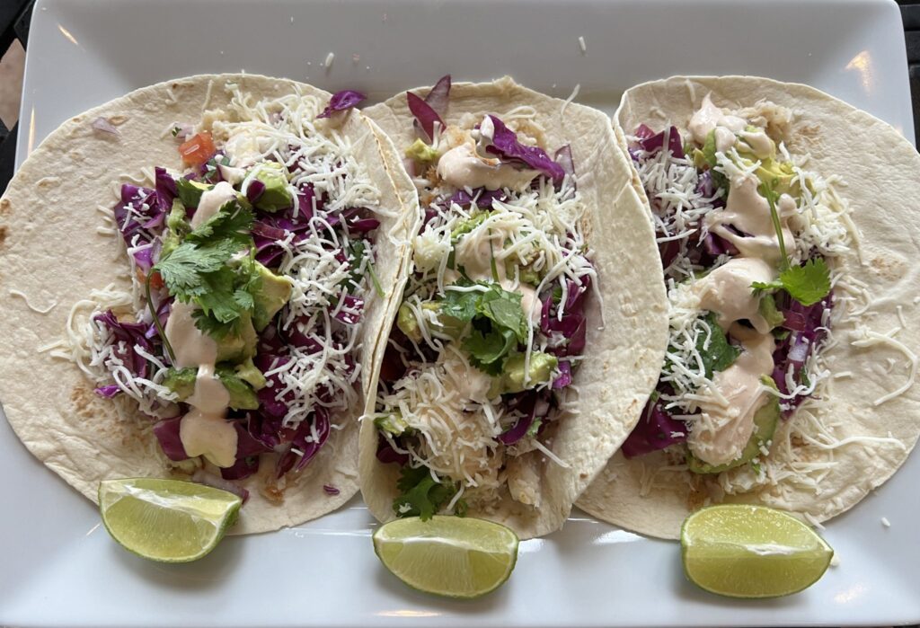 Fish tacos with flaky white fish, shredded cabbage, tomato, sauce, and shredded cheese sit on a plate with three lime wedges. Rubio's