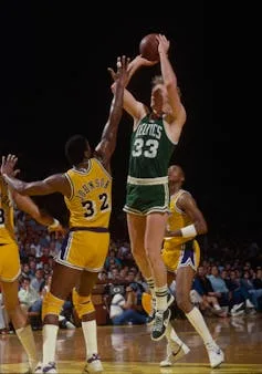 Man in green jersey shoots a basketball over the outstretched hand of man in yellow jersey.