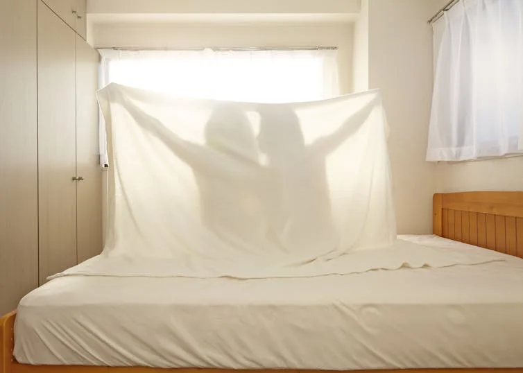 Silhouettes of two women sitting on a bed holding up a white sheet.