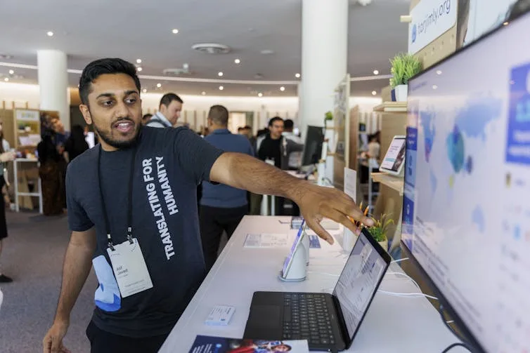 A man in a blue t-shirt points to the screen of a laptop opened on a white table in front of a large poster.