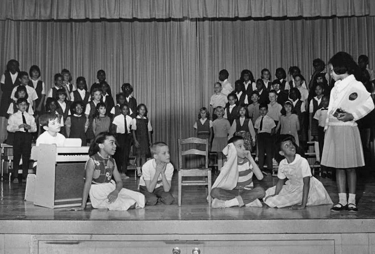 A black and white photo of children on stage, many of them standing on chorus risers.