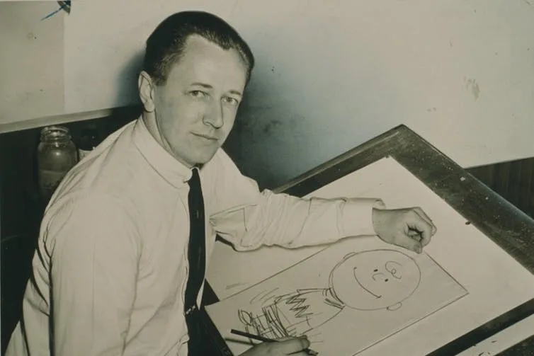 A black and white photo of a man in a white suit and dark tie, seated at a drawing table with a cartoon of a boy in front of him.