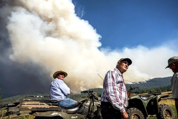 Two older men on ATVs watch the sky as a cloud of smoke rises behind them.