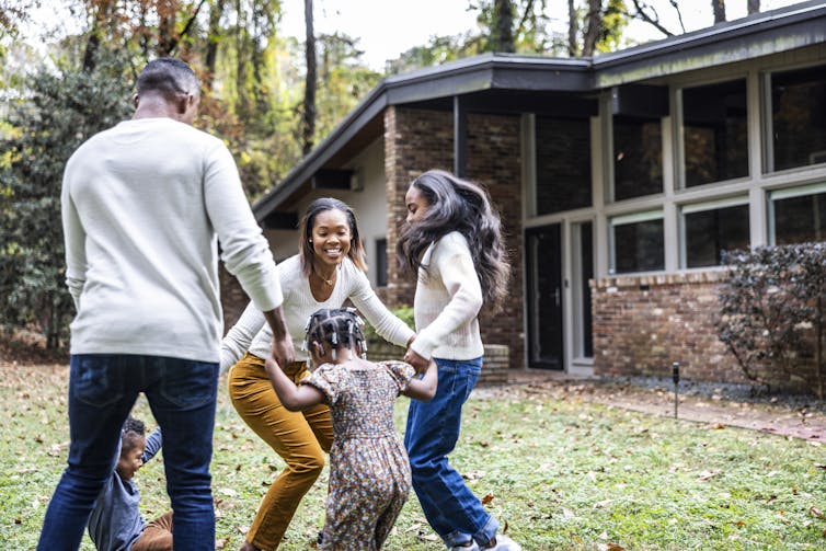 A Black family plays with young children in front of a suburban house.