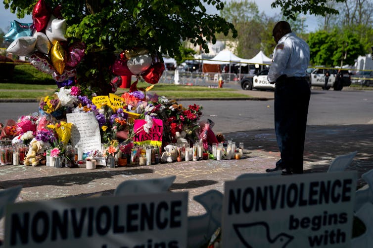 Person gazing at makeshift memorial at the foot of a tree, police cars in the background, and nonviolence signs in the foreground