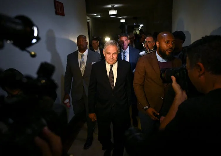 Light shines on white man with grey hair walking with group of men in suits down a corridor while cameras film them