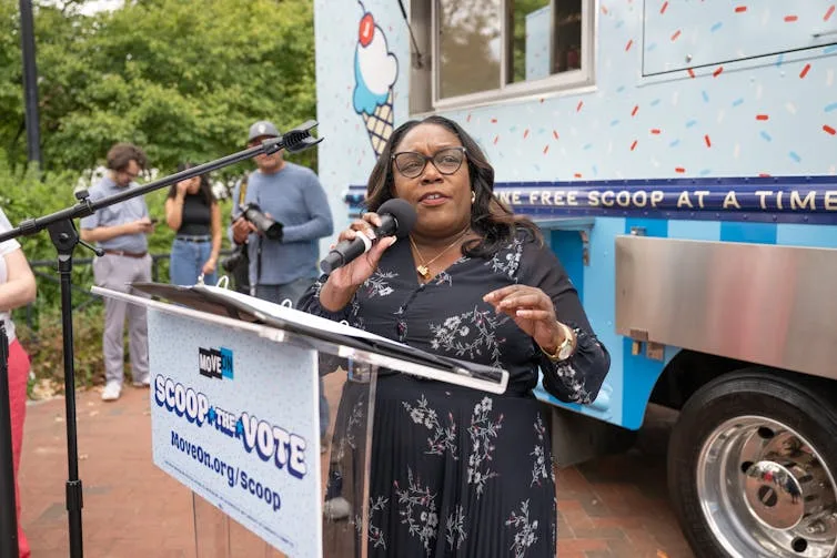 Woman holds microphone while speaking at a podium in front of an ice cream truck