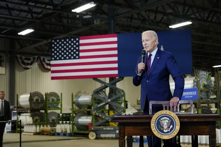 Biden talks into mic in a factory with big American flag in background