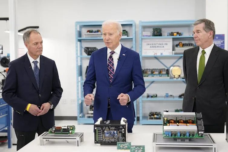 Biden stands at table that holds examples of technology, flanked by two other men