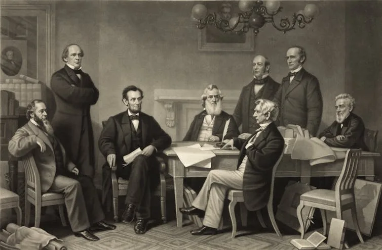 A group of formally dressed men gather around a small table with a piece of paper on it.