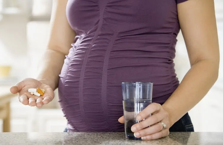 Torso of a pregnant woman holding a glass of water in one hand and vitamins in the other.