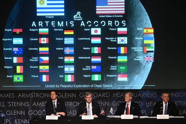 Four officials sitting at a table in front of a screen with the flags of countries party to the Artemis Accords.