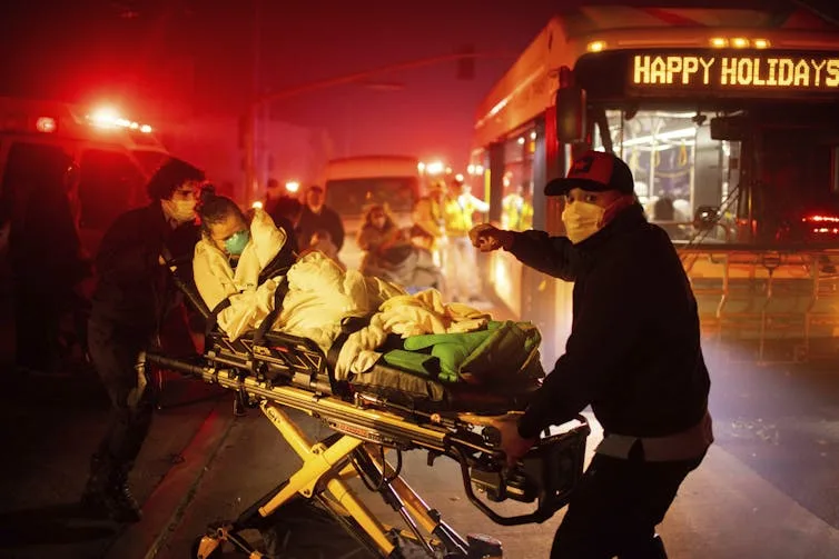 Emergency responders push a person laying on an ambulance stretcher with smoky air and a city bus nearby.