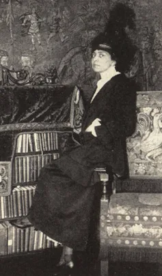 Woman wearing a large, plumed hat, seated on the arm of a chair next to a bookshelf.