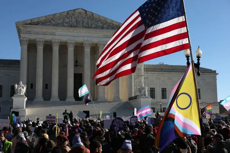 A crowd of protesters holding signs and flags before the steps of a majestic, columned, white building.