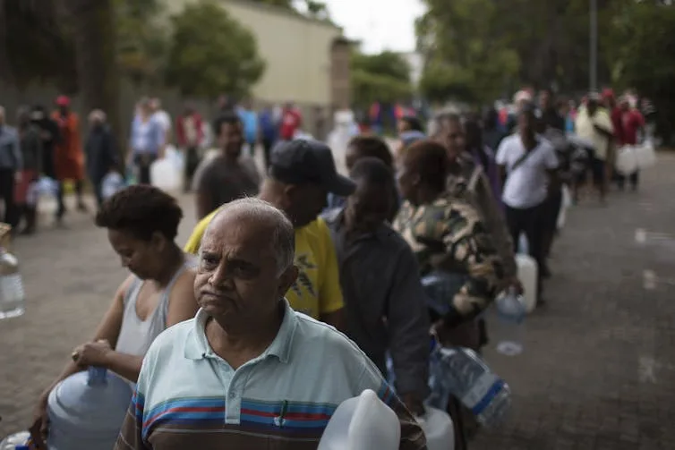 People holding jugs of all kinds stand in a long line for water.