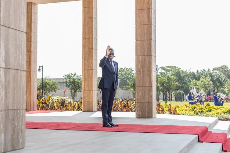 A man in a suit waving from a terrace and standing on a red carpet.