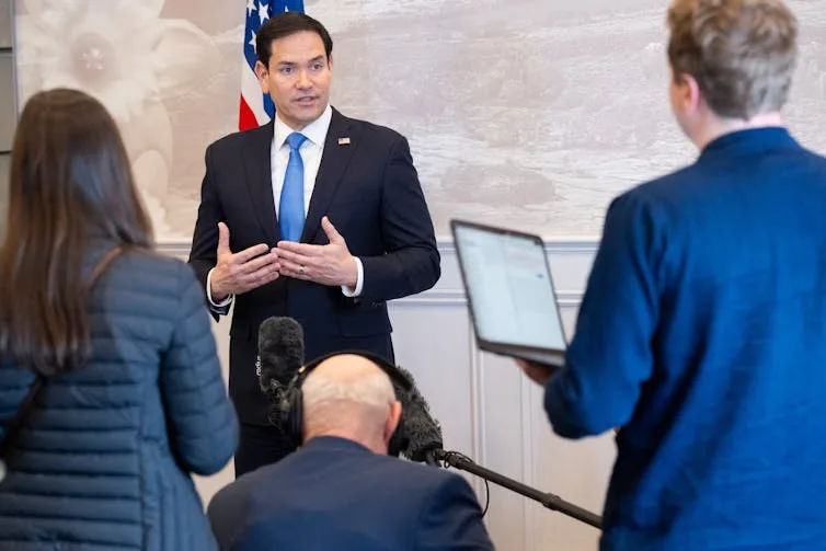 A man with dark hair and a black suit gestures facing people looking at him.