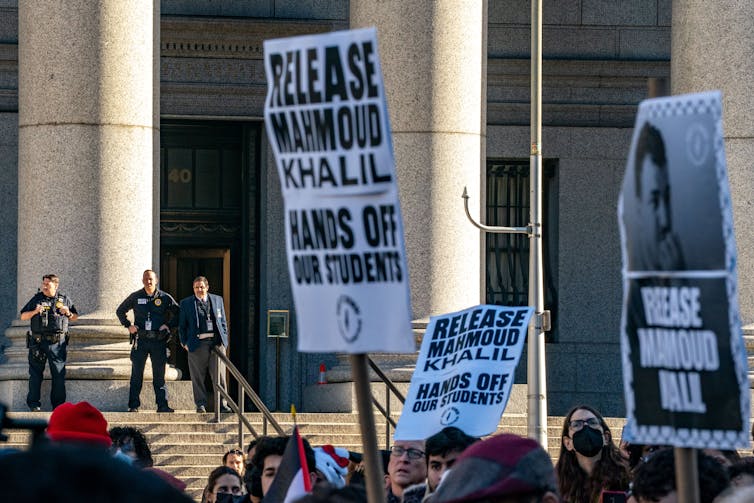 People stand together and hold signs that say 'Release Mahmoud Khalil' in front of a large building while police officers in black watch.